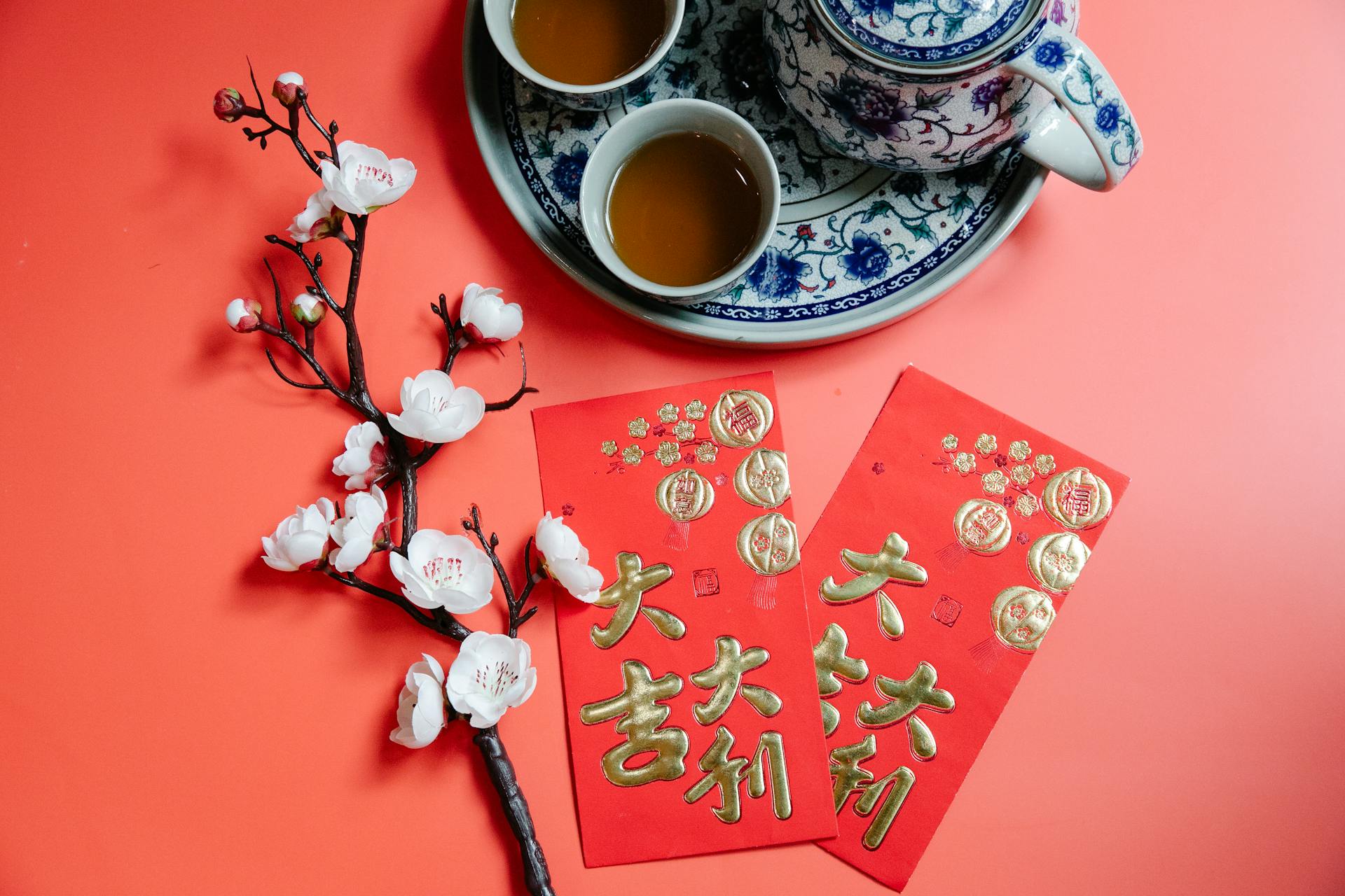 Top view of oriental packets with hieroglyphs against blossoming flower sprig and tea set during New Year holiday on red background