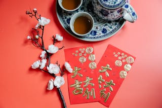 Top view of oriental packets with hieroglyphs against blossoming flower sprig and tea set during New Year holiday on red background