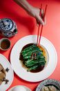 From above of crop unrecognizable person with chopsticks above tasty pak choi in soybean sauce against tea on red background