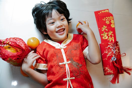Top view of cheerful ethnic child with fresh oranges and red packets looking up while lying on floor during New Year holiday
