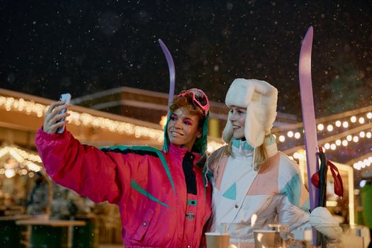 Two women in colorful ski gear take a selfie at a festive outdoor ski resort at night.