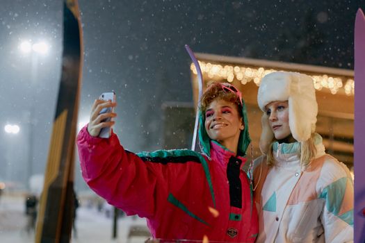 Two friends pose for a selfie in colorful ski jackets during a snowy winter evening.