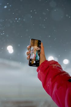 Woman in red jacket taking a selfie with smartphone outdoors in the snow at night, capturing a winter moment.