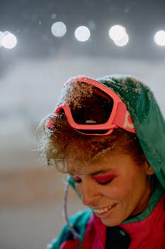 Close-up of a woman smiling in winter snow wearing ski goggles and a jacket, enjoying the snow fall.