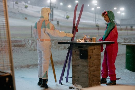 Two skiers in colorful ski suits warm up by an outdoor fire on a snowy winter night.