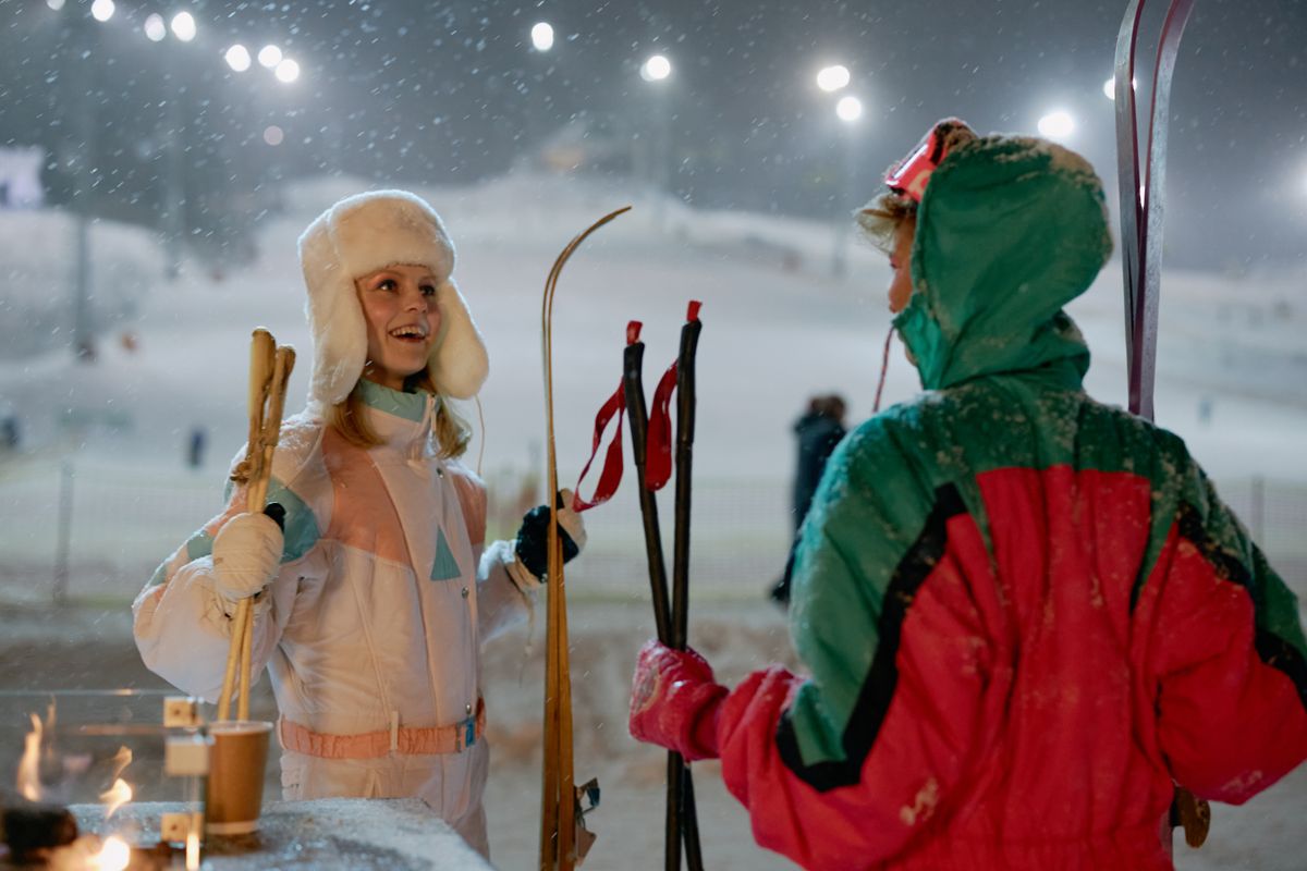 Two women in winter clothing holding skis and drinks at a snowy resort.