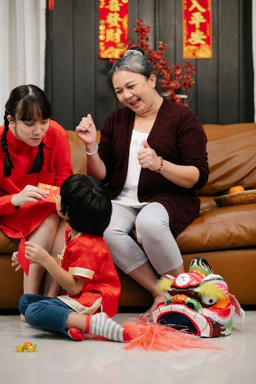 Cheerful ethnic female teenager with red envelope talking to unrecognizable brother against grandma during New Year holiday at home