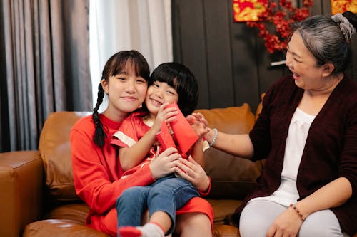 Smiling ethnic female adolescent embracing sibling with red pocket against grandma on couch during New Year holiday at home