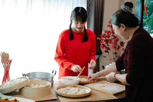Free Smiling ethnic grandma with female teen preparing Chinese dumplings while talking at table with traditional steamers in house Stock Photo