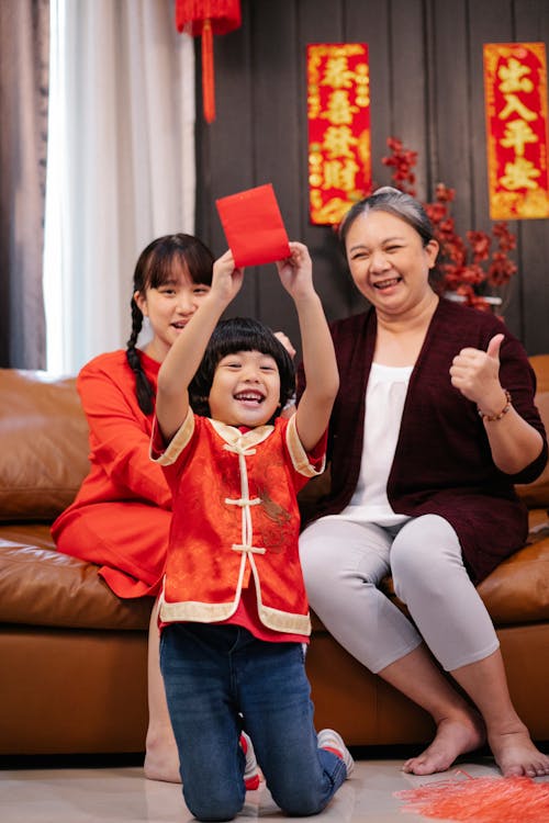 Excited ethnic child with red packet against female teenager and grandmother celebrating New Year holiday in house room