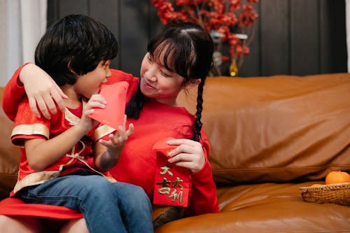 Content ethnic female teenager embracing sibling with red envelope while looking at each other during festive event on sofa