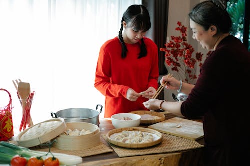 Free Cheerful ethnic grandma with attentive teen preparing dumplings at table with traditional steamers during New Year holiday at home Stock Photo