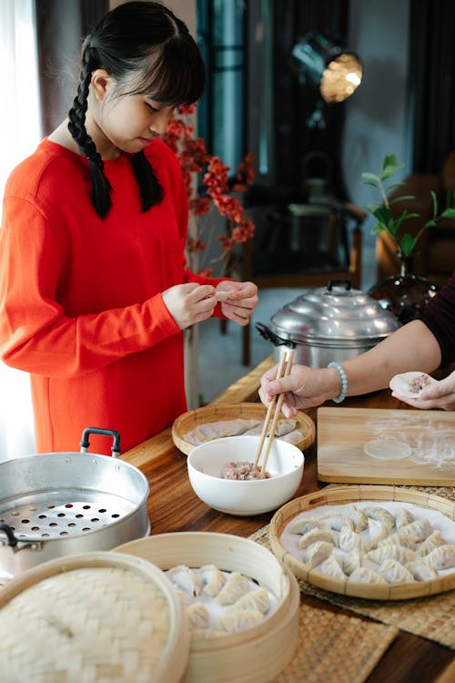 Free Ethnic female teenager and crop unrecognizable woman with chopsticks preparing dumplings with ground meat filling at home Stock Photo