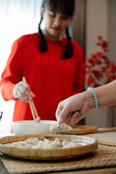 Crop Asian teenage girl with dark hair using chopstick while helping unrecognizable mother to prepare traditional Chinese jiaozi dumplings