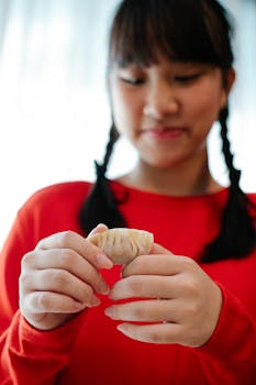 Crop happy ethnic female teenager with braids in casual clothes smiling while folding traditional Chinese jiaozi in kitchen
