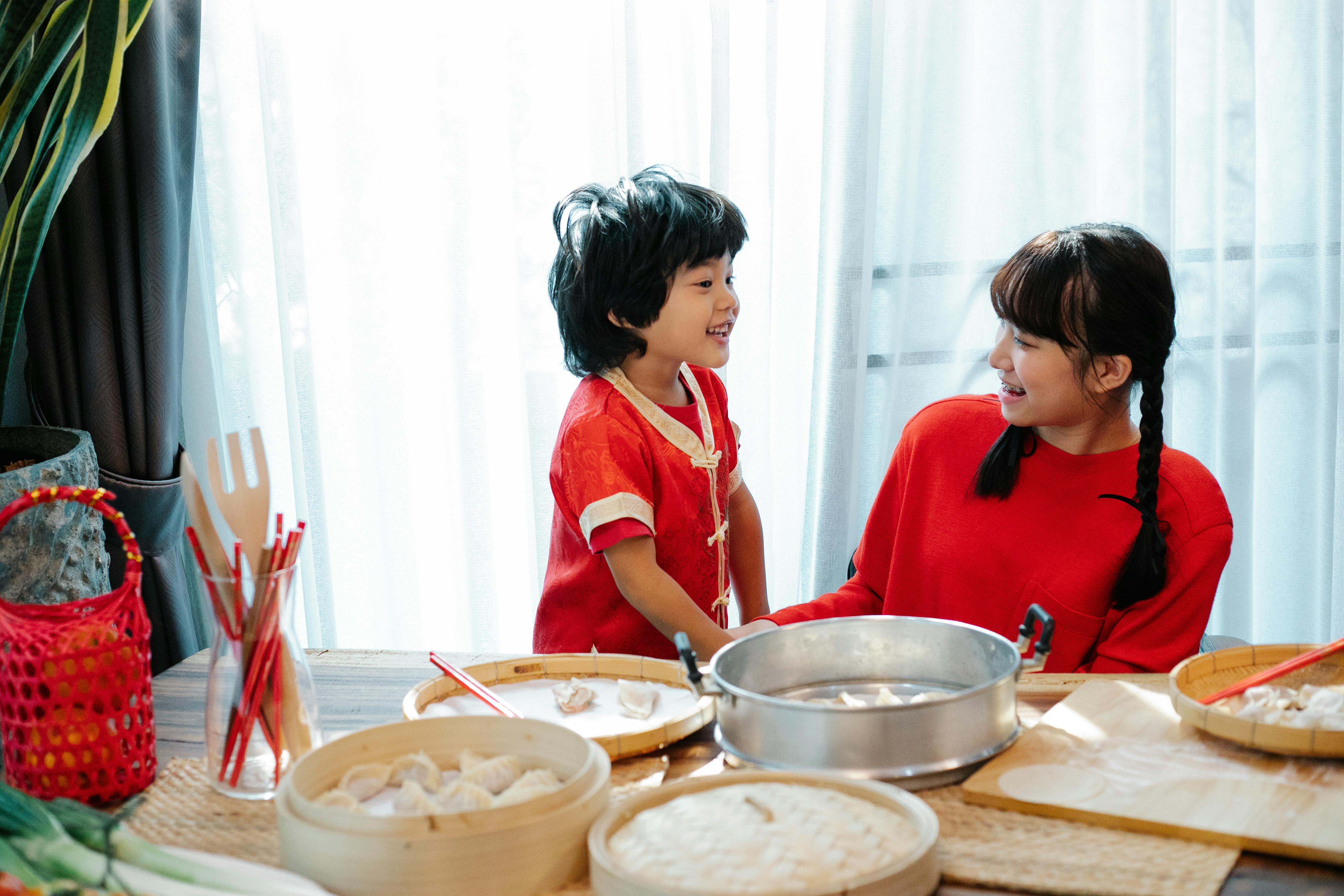 Positive Asian boy and teen sister smiling happily while cooking homemade jiaozi dumplings at table together