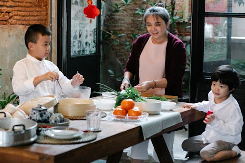 Ethnic grandmother preparing ingredients for cooking traditional lunch while cooking with boys at table in kitchen