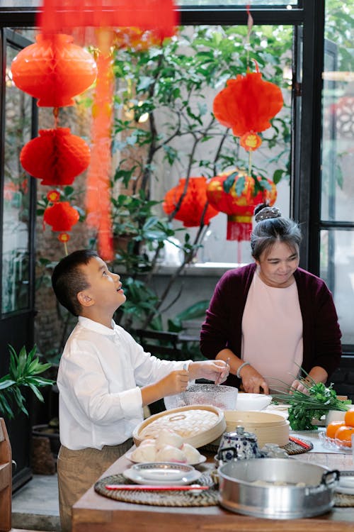 Amazed ethnic boy standing near grandmother cutting vegetables while making ground meat for dinner in kitchen with Chinese lanterns