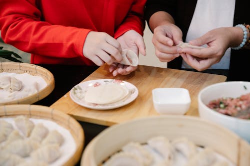 Crop anonymous teen girl making traditional Chinese jiaozi dumplings while cooking lunch together at table with ingredients and bamboo steamer