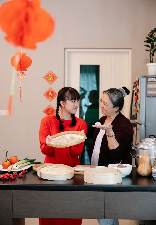 Free Smiling ethnic old woman demonstrating Chinese jiaozi dumpling to granddaughter standing with bamboo tray in room with oriental lanterns Stock Photo