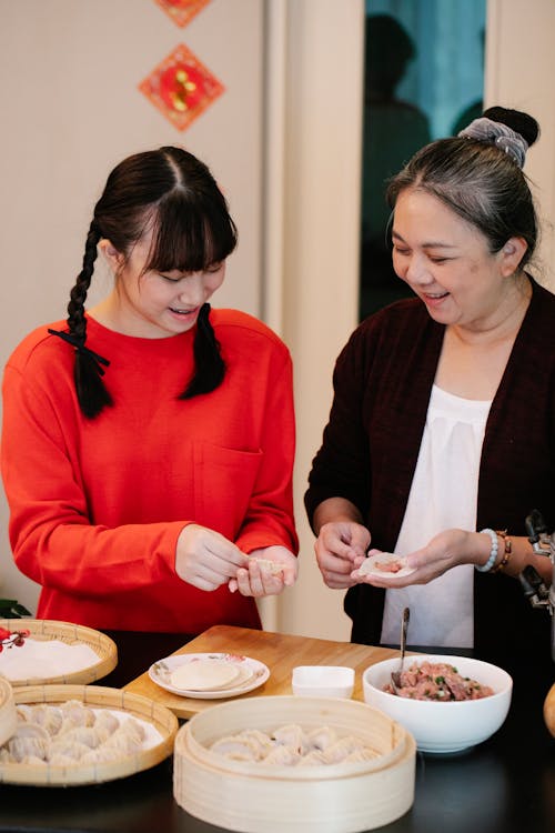 Positive ethnic teen granddaughter helping grandmother making Chinese jiaozi dumplings with dough and minced meat in kitchen