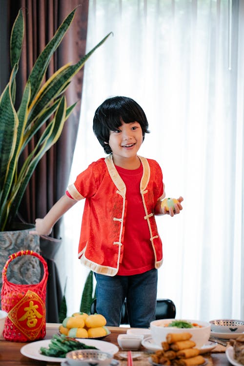 Cheerful ethnic child with rice bun looking away at table with assorted dishes and lantern during New Year holiday