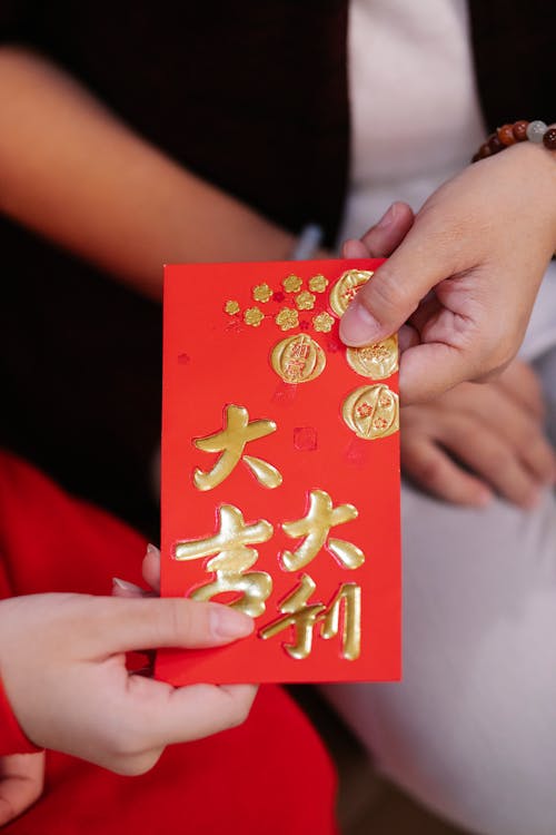 Crop unrecognizable female passing red pocket with hieroglyphs and bauble ornament to relative during New Year holiday