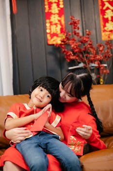 Ethnic female teenager embracing smiling sibling with red packet while interacting on couch during New Year holiday at home