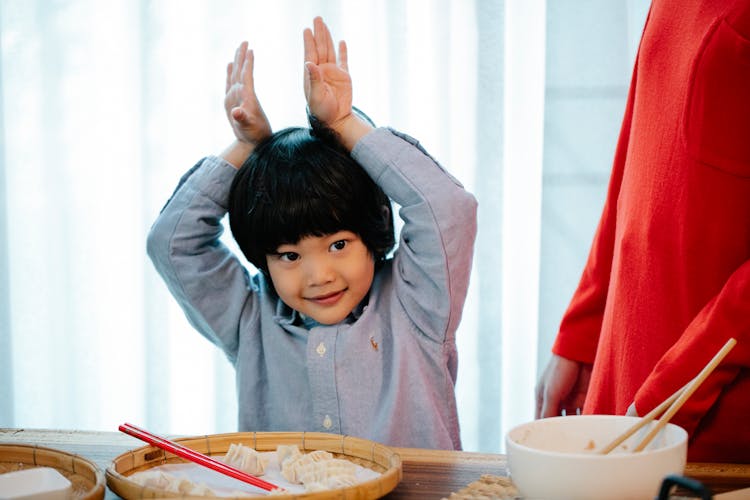 Positive Asian Kid Making Bunny Ears With Hands With Hands In Kitchen