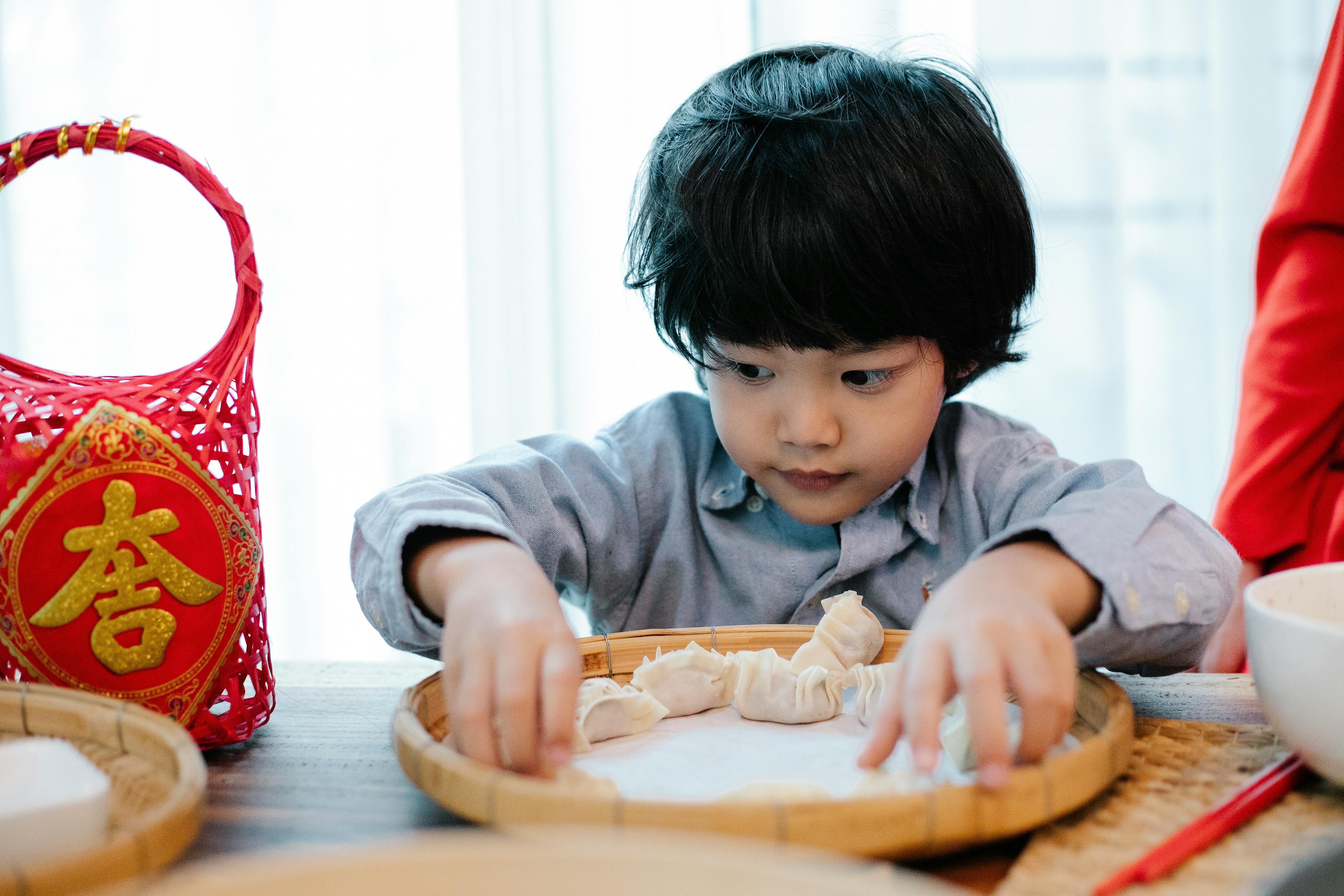 A cute child carefully prepares dumplings indoors, with a red basket adding a cultural touch.