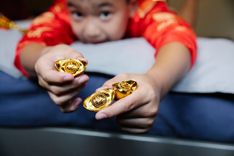 Positive Little Chinese Kid Lying On Bed And Showing Traditional Gold Ingots