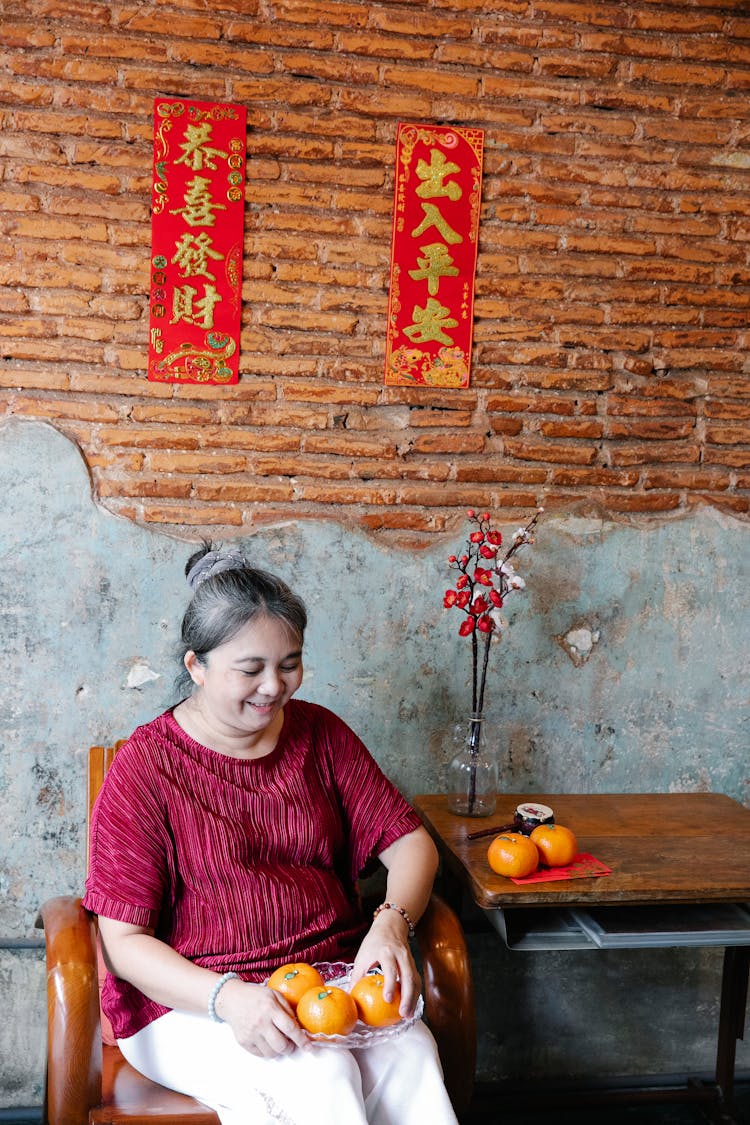 Smiling Mature Ethnic Woman Serving Fresh Fruits On Plate Sitting On Chair