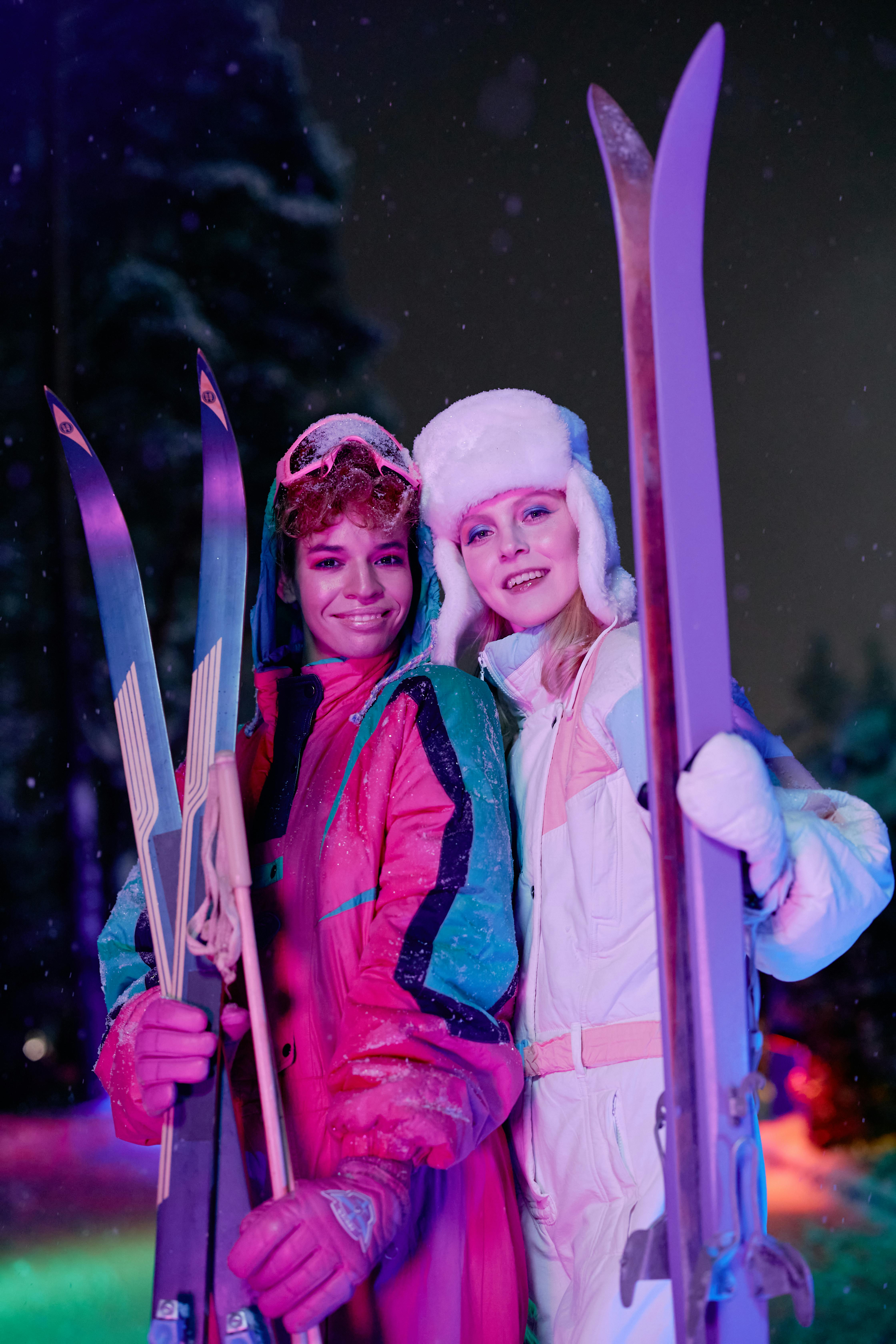 Prescription Goggle Inserts - Two women enjoying a colorful nighttime skiing adventure in winter attire, holding skis under vivid lights.