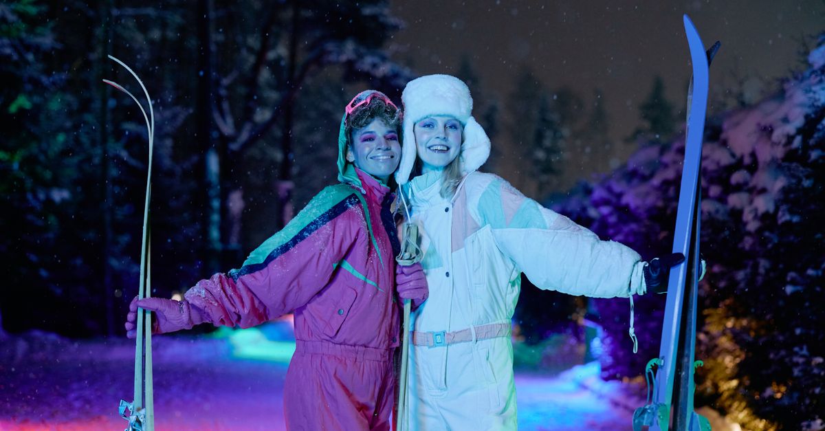 Two women enjoy night skiing under vibrant neon lights, showcasing stylish winter fashion.