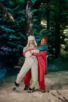 Two women in winter gear smiling and hugging in a snowy forest at night.