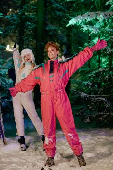 Two women in vibrant ski outfits pose joyfully in a snowy forest at night.