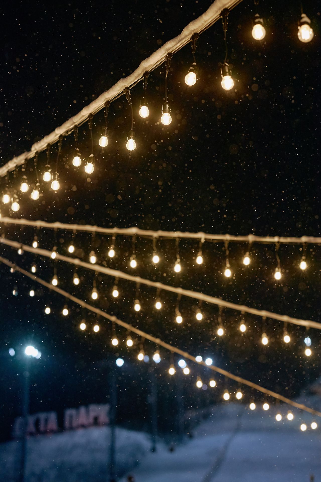 Warm string lights glowing over fresh snow at night as snowflakes fall.