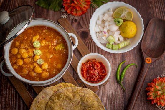 Pot of chickpea curry with scallion rings beside puris, onions, lemon and red chutney