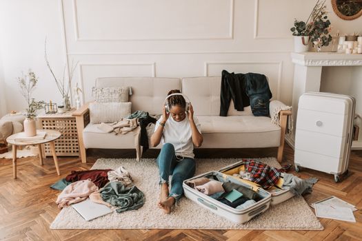 A woman packs a suitcase in her living room while listening to music, surrounded by clothes and a cozy atmosphere.