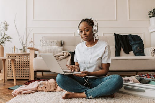 Woman sitting on the floor with a laptop and headphones, preparing to pack with clothes around.