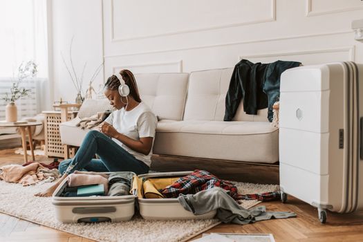 A young woman in a living room packs a suitcase with headphones on, preparing for a trip.