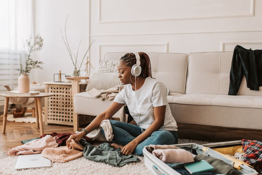A woman with braided hair packs a suitcase at home, listening to music with headphones.