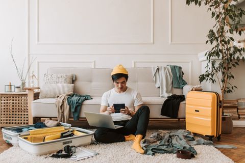 A young man uses multiple gadgets while sitting in a stylish, cozy living room with a yellow suitcase and open luggage.