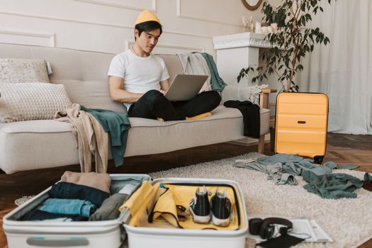A young man in yellow accessories works on a laptop among unpacked luggage at home.