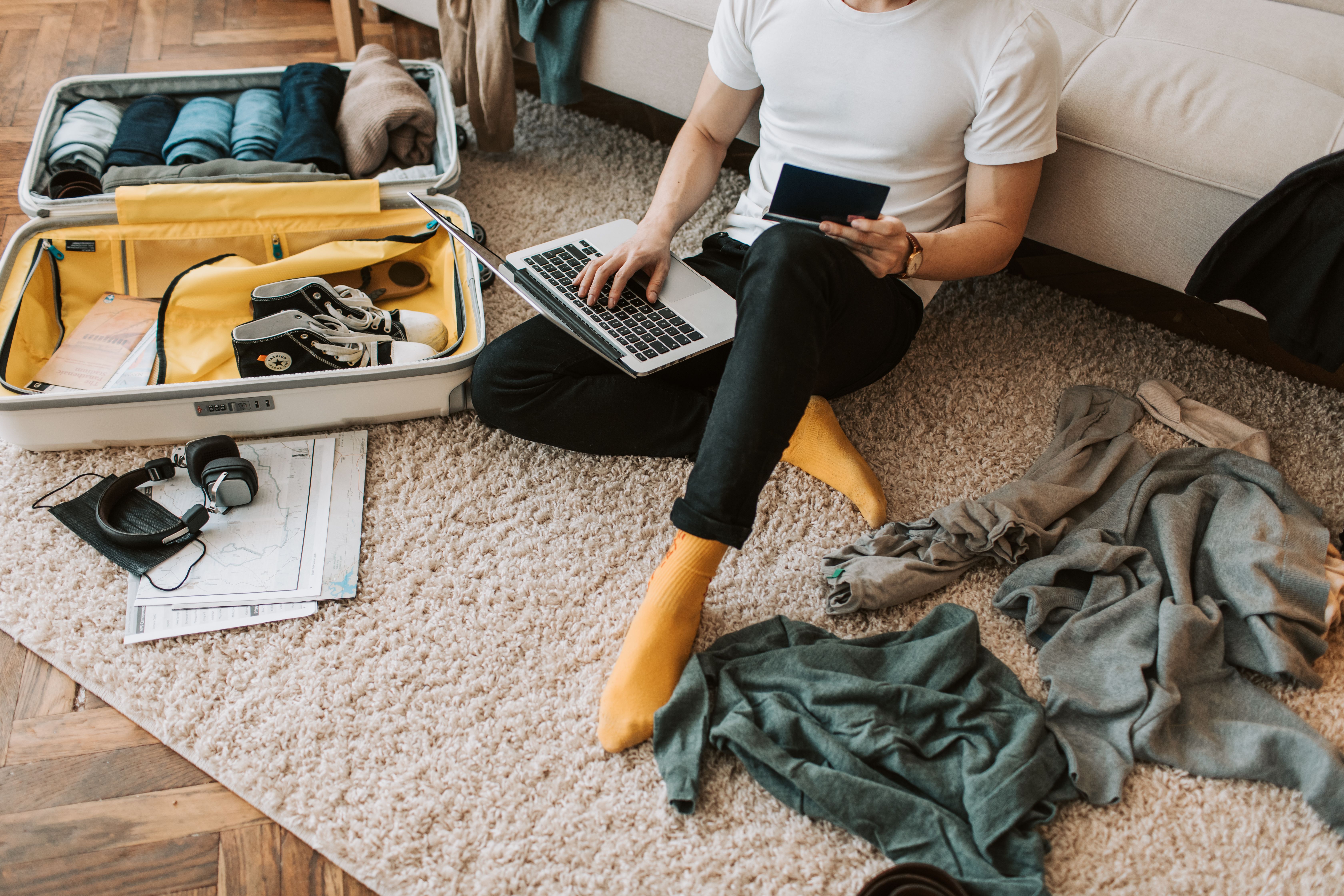 Traveller packs a suitcase while using a laptop and holding a passport in a hotel room