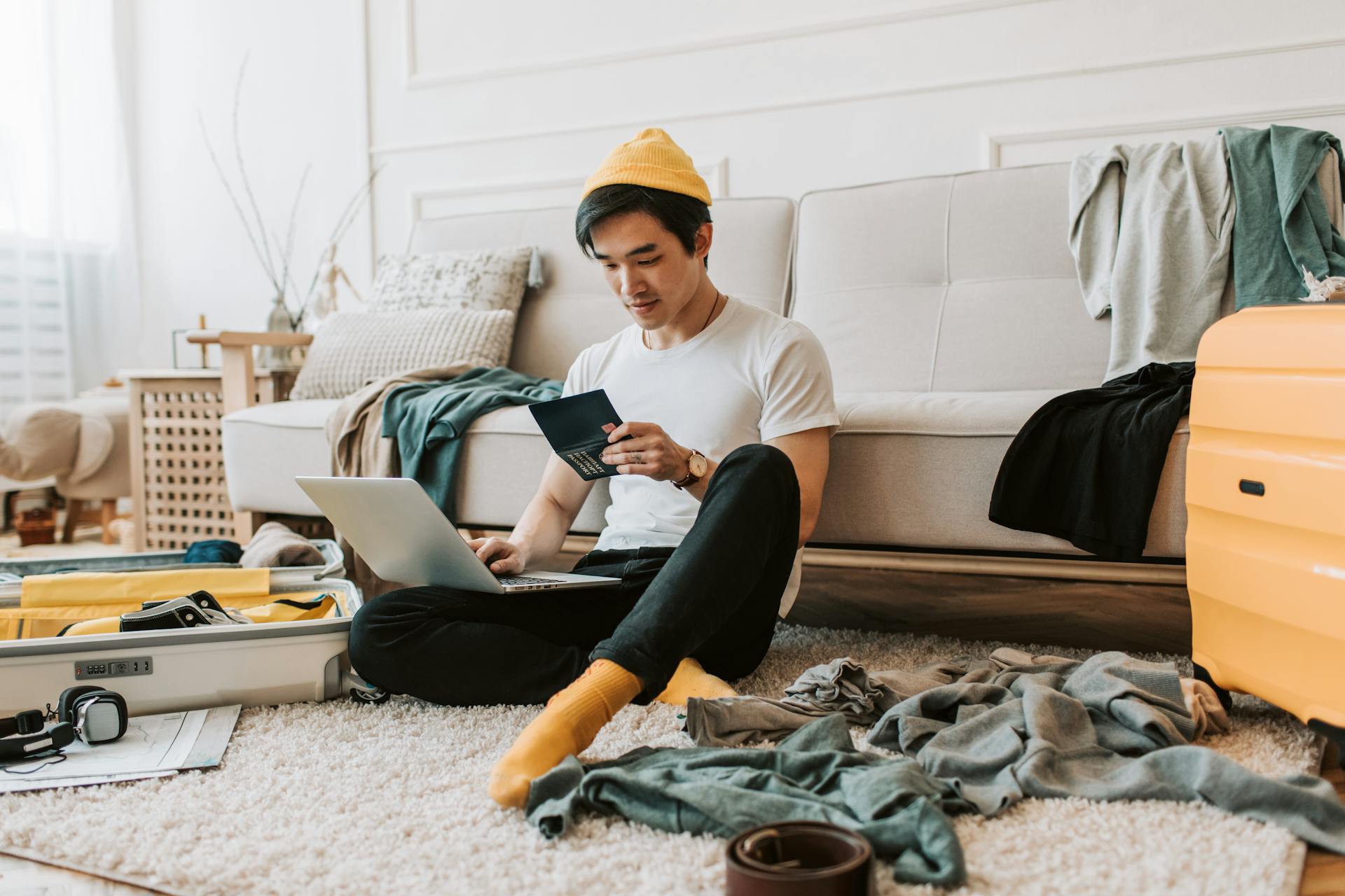 A young man in a messy living room preparing for travel with a laptop and passport.