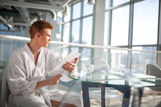 A man in a bathrobe sits pensively by a glass table inside a modern building.