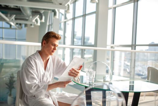 Man in bathrobe reading menu indoors, surrounded by natural light and glass décor.
