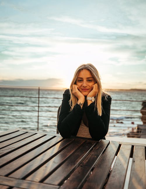 Portrait of a Blonde Woman Leaning against Wooden Table and Sea in Background