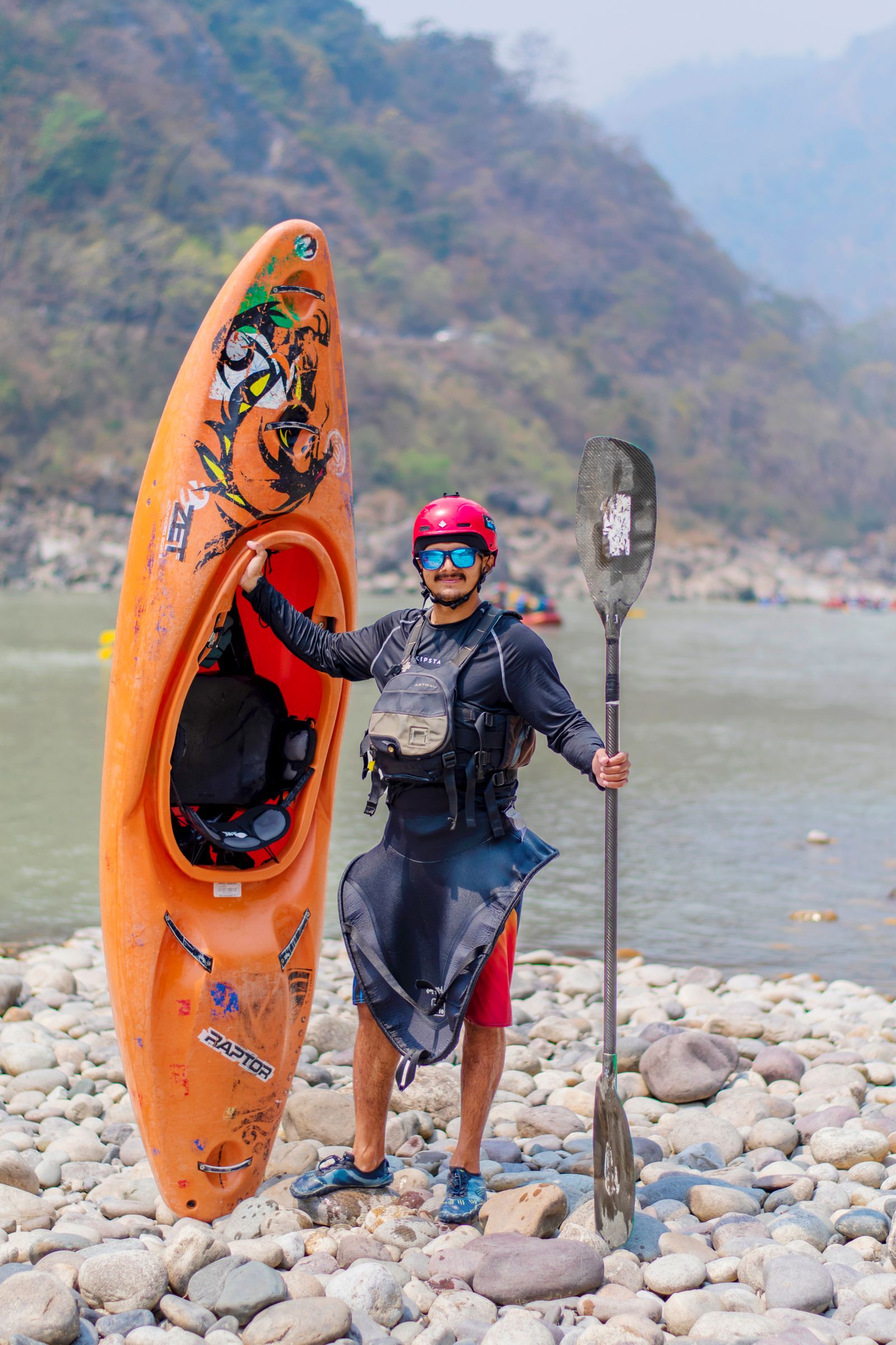 Person standing beside the water holding a kayak and paddle while preparing outdoor water-sports equipment